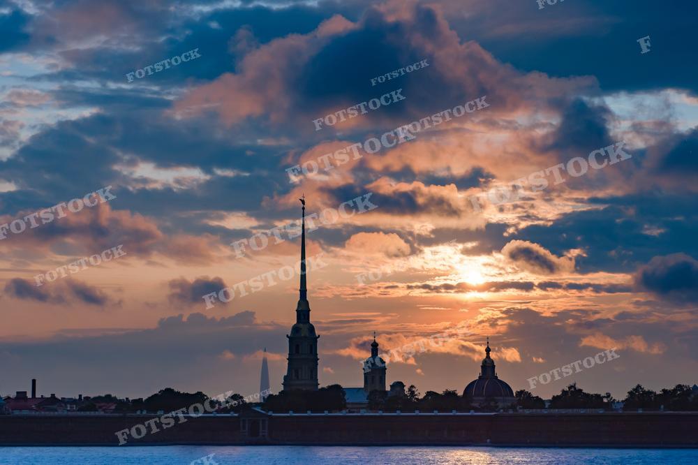 На этой фотографии мы видим захватывающий вечерний вид на Петропавловскую крепость в Санкт-Петербурге. Солнце скрывается за облаками, окрашивая небо в тёплые оттенки оранжевого и голубого, что создаёт контраст с темной силуэтной линией крепости. Силуэты шпилей и куполов крепости вырисовываются на фоне заходящего солнца, подчёркивая историческую и культурную значимость этой достопримечательности. Фотография идеально подойдёт для статей о Санкт-Петербурге, для использования в учебных материалах по истории России, или как элемент дизайна в тематических публикациях, связанных с культурным наследием.